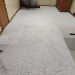 Light-colored carpeted basement room with visible rectangular patch in the center. Some furniture is against the walls.