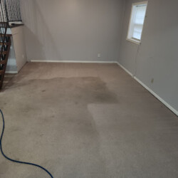 A carpeted room with noticeable stains, a staircase on the left, and a window on the right wall.