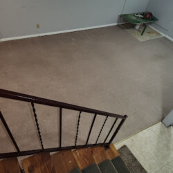 View from staircase showing a mostly empty carpeted room, with a small table and some items in the far corner.