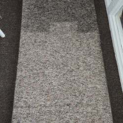 A rectangular piece of carpet with a darker, damp section on one end placed on a brown floor.