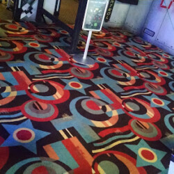 Colorful carpet with bold geometric patterns in a public indoor space, near signage and a laser tag entrance.