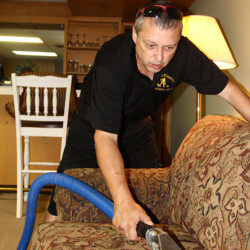 A man uses a vacuum extraction tool to clean a patterned sofa in a living room.