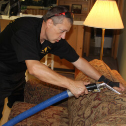 A man uses a professional cleaning tool to clean a patterned sofa in a living room.