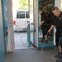 A man uses a cleaning sprayer on a carpeted floor near a glass door inside a room with cleaning supplies.
