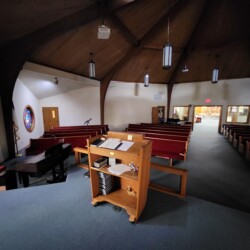 View from the pulpit inside a church sanctuary with pews, piano, and stained glass window visible.