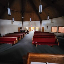 Interior of a church with empty red pews, wooden ceiling, and lights hanging from above; view from the altar.