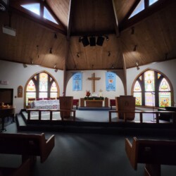 Interior of a church sanctuary with stained glass windows, altar, wooden ceiling, and pews visible.