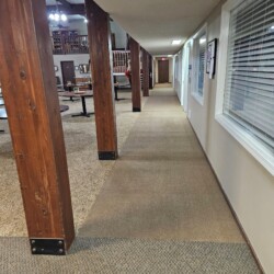 Long hallway with wooden support beams, carpeted floor, and windows and doors along the right side.