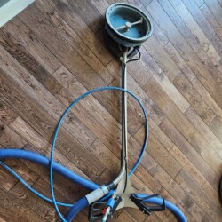A floor cleaning machine with attached blue hoses is positioned on a wooden floor near a wall.