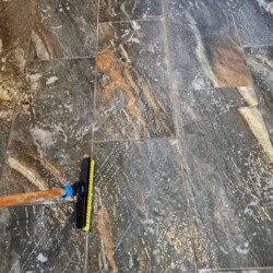 A mop with a yellow brush is cleaning a tiled floor with soap and water.