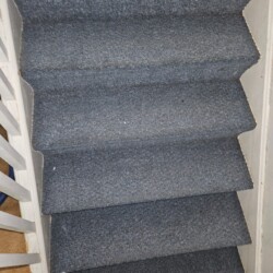 A set of steep, narrow stairs covered with blue-grey carpet, seen from above. White railings are on the left side.
