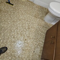 A bathroom floor with tile pattern is wet, showing water pooling near a toilet and a broom leaning against the wall.