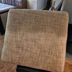 A square, brown upholstered seat cushion shown in close-up indoors, with a faint horizontal line across the fabric.