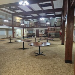 Large, carpeted room with round tables, wooden beams, an American flag, and a second-floor balcony.